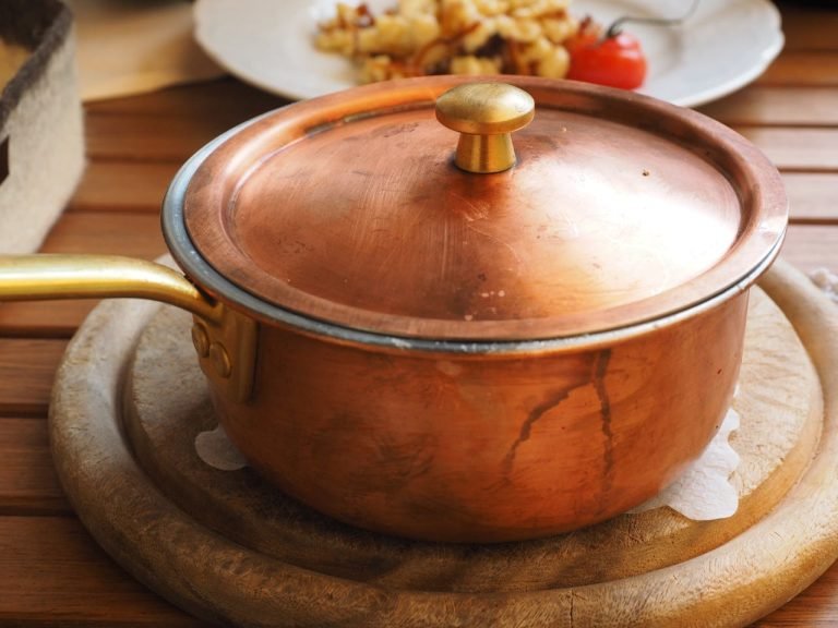 A rustic copper pot sits on a wooden table with a background of kitchenware.